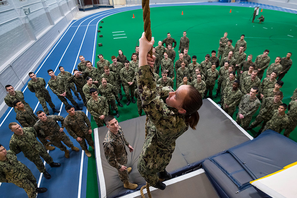 NROTC midshipmen climbs a rope with a crowd of fellow midshipmen looking on