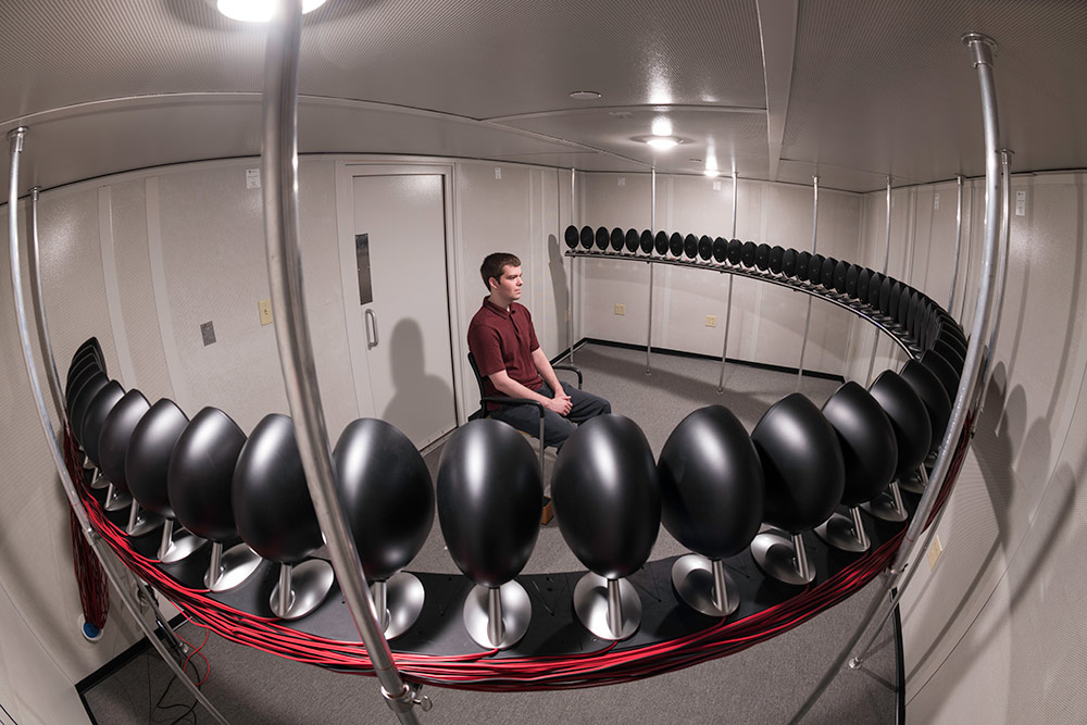 graduate student sits in a sound booth in the center of a circle of speakers