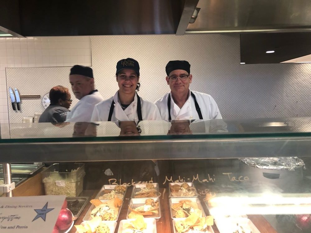 Feature image two officers in chefs uniforms smile from a kitchen window.