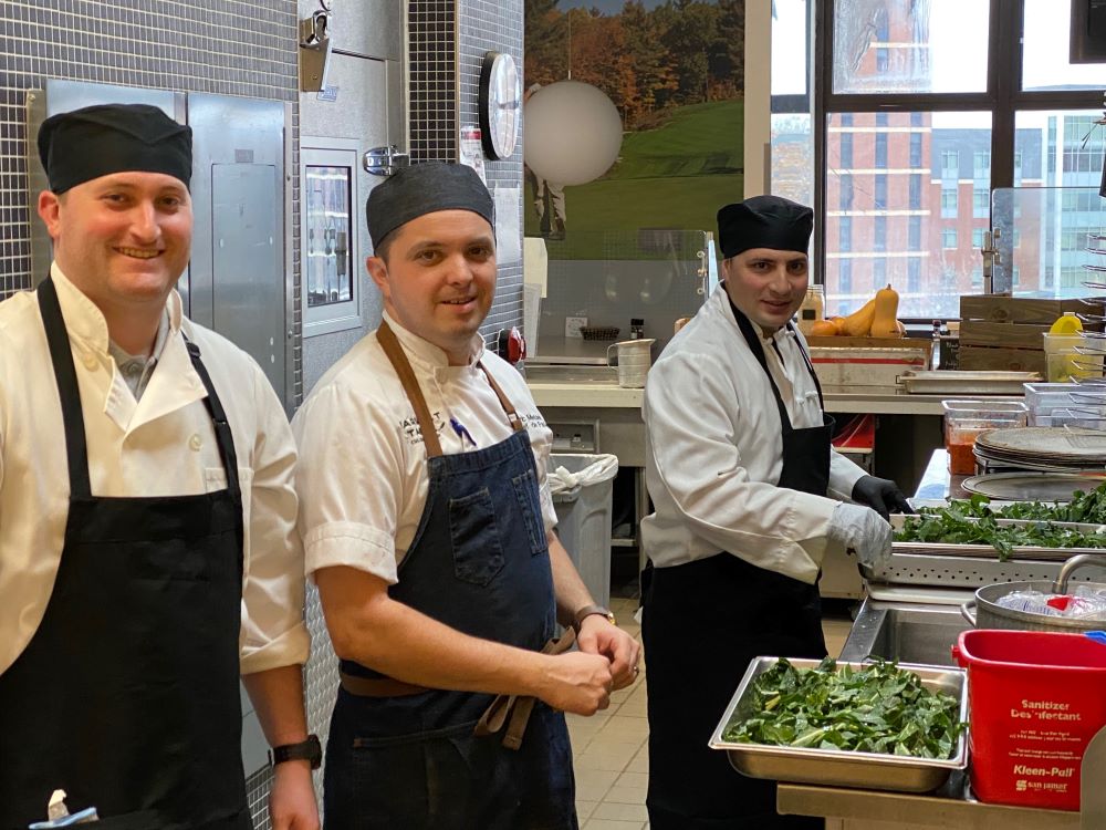 Three people in chefs uniforms prepare greens in a kitchen.