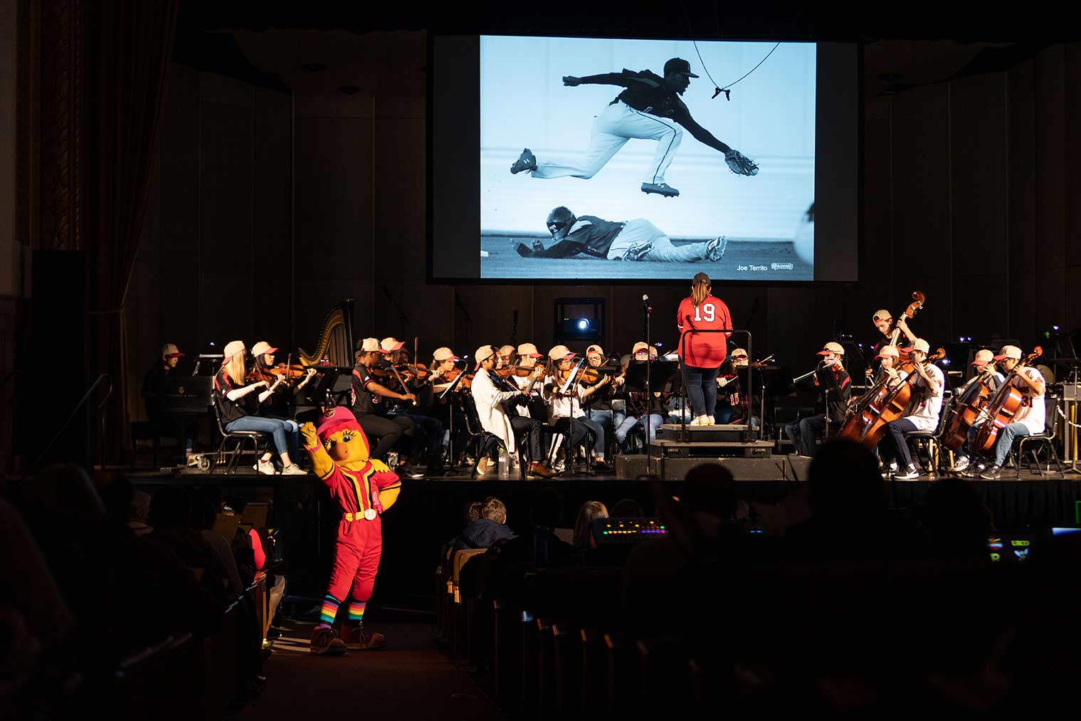 Feature image orchestra musicians on stage, with conductor's back facing the audience, all musicians in baseball uniforms, with the Red Wings baseball mascot in the foreground and a screen in the background showing Roberto Clemente playing baseball.