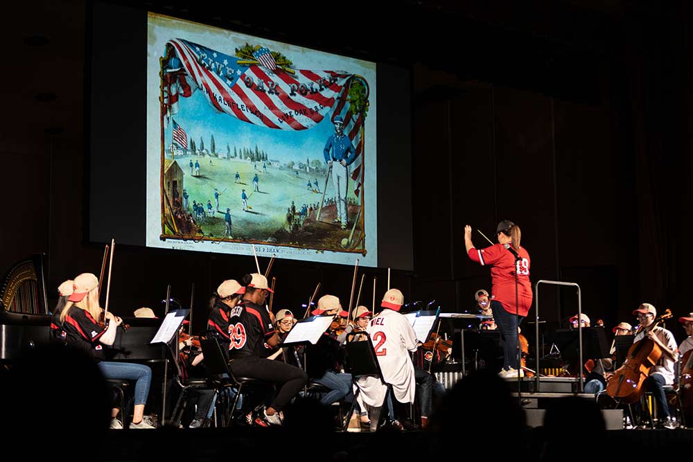 conductor and orchestra musicians perform in baseball uniforms, with an historic image of a baseball game and American flag on the screen behind them.