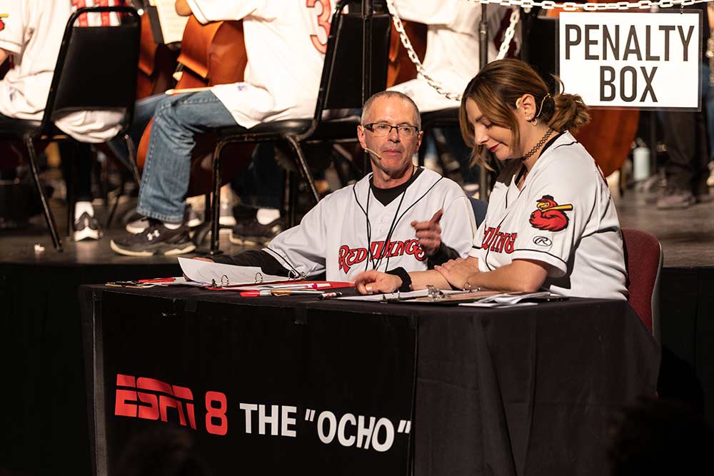 two radio announcer sit at a table in front of a stage, with the words PENALTY BOX and ESPN 8 The Ocho.