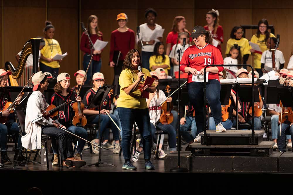 woman speaks into a microphone on stage in front of orchestra musicians wearing baseball uniforms.