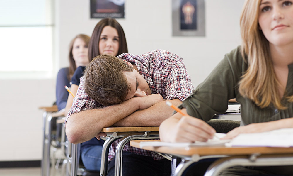 a classroom, where a teen sleeps with his head down on his desk