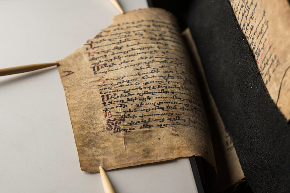 single handwritten page of an ancient book being held flat on a scanner by wooden knitting needles.