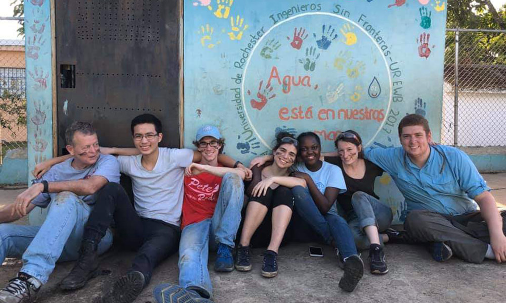Feature image group portrait of students leaning against a school wall painted with children's handprints.