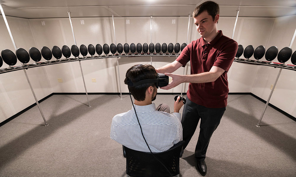 person sitting, wearing a virtual reality headset, and surrounded by a circle of audio speakers