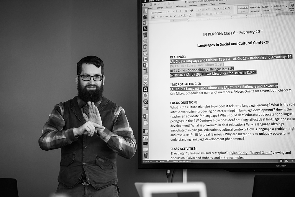 student speaking American Sign Language in front of a large screen in a classroom.