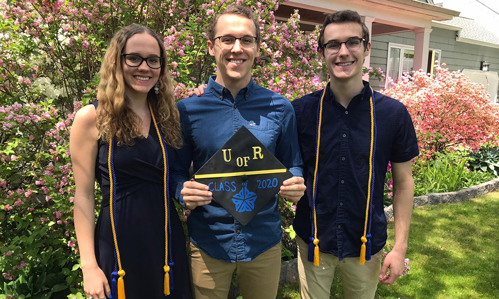 Feature image triplets in commencement regalia pose and hold mortarboards outside their home.