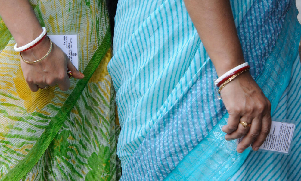 Close up of the bodies and arms of two women in saris wearing bracelets and holding electoral commission identity cards.