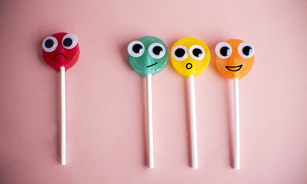 Four lollipops with cartoon eyes and facial expressions on pink background.