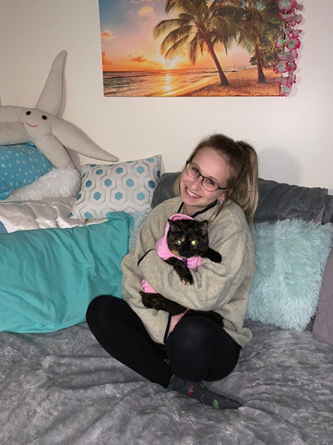 Student on bed, holding cat, in dorm room.
