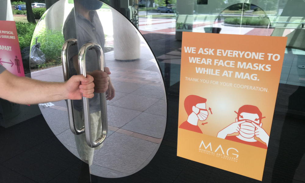 Feature image Man's arm opens the doors of the Memorial Art Gallery, which feature a bright orange sign asking everyone to wear a mask while at MAG>