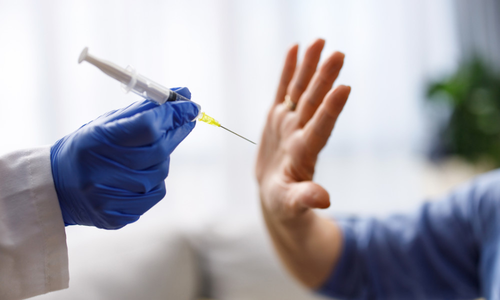 Close-up of a health professional's gloved hand holding a syringe while an unrecognizable person has their palm out to reject the vaccination.