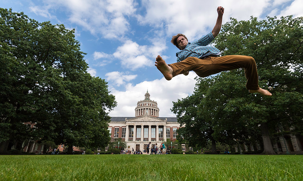 Feature image student does a leap on the quad that makes it look like he is jumping over Rush Rhees Library.