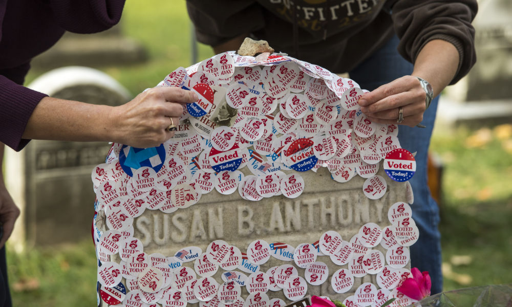 Hands place "I voted today" stickers on Susan B. Anthony's headstone.