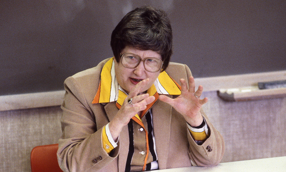 Feature image archival photo of Mary Young, seating as desk in front of a blackboard, speaking.
