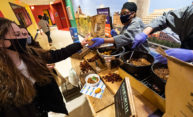 cook from Dining Services hands a sample of chili from a large pot to students on the other side of the table, all are wearing masks.
