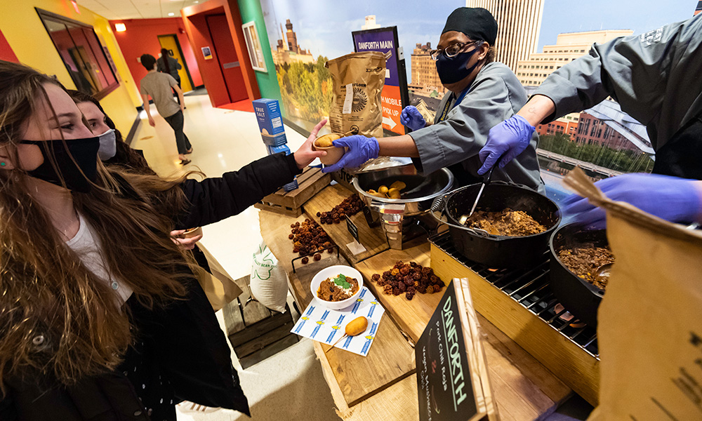 Feature image cook from Dining Services hands a sample of chili from a large pot to students on the other side of the table, all are wearing masks.