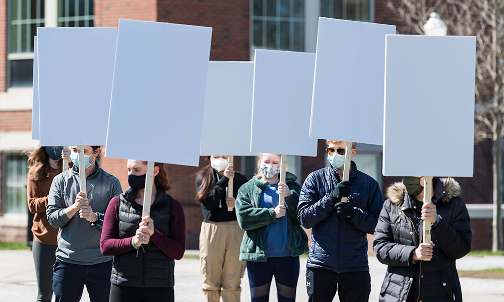 a row of students stands outside on the quad, each holding a blank protest sign.