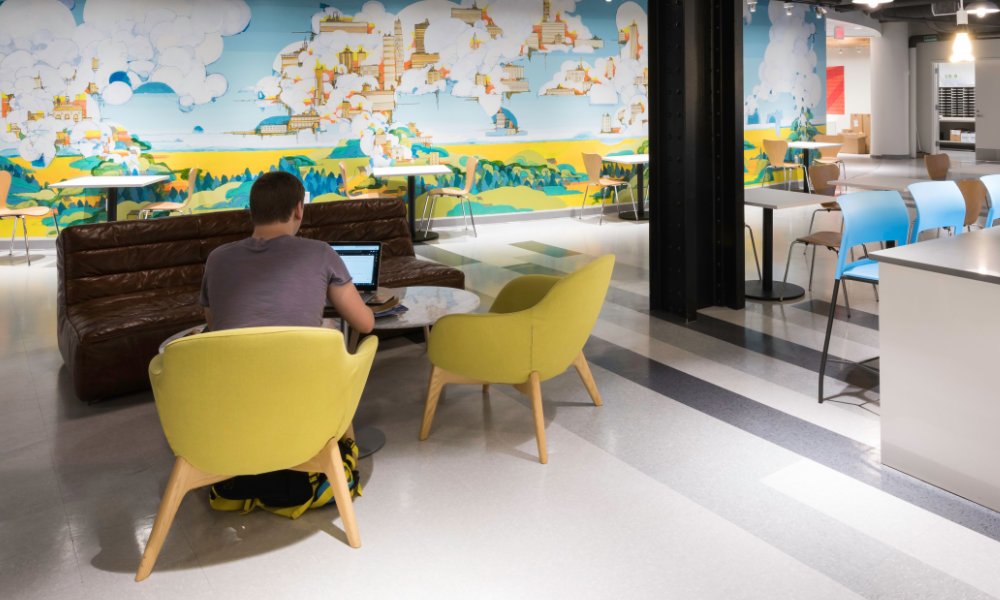 Seen from behind, a man sits in a yellow chair and looks at his laptop in the NextCorps business incubation space at Sibley Square, complete with colorful mural and modern shared workspaces.