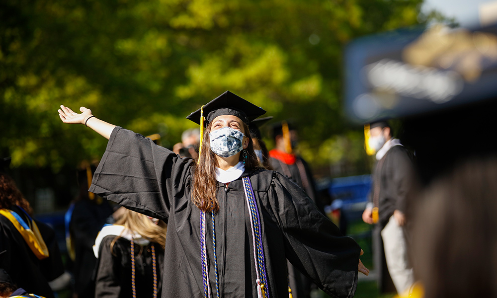 student in a cap and gown throws there hands up in the eye and smiles.