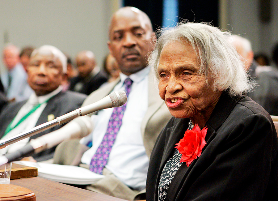 Olivia Hooker in the foreground, seated at a table and speaking into a microphone.