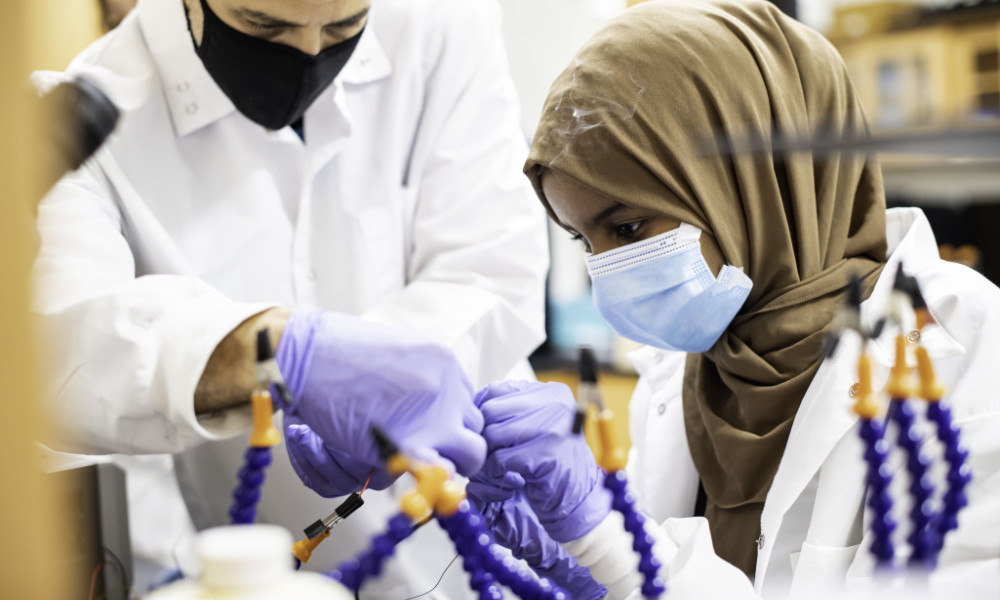 High school student in a head scarf, face mask, and lab gear practices soldering to repair lab equipment while a lab tech looks on.