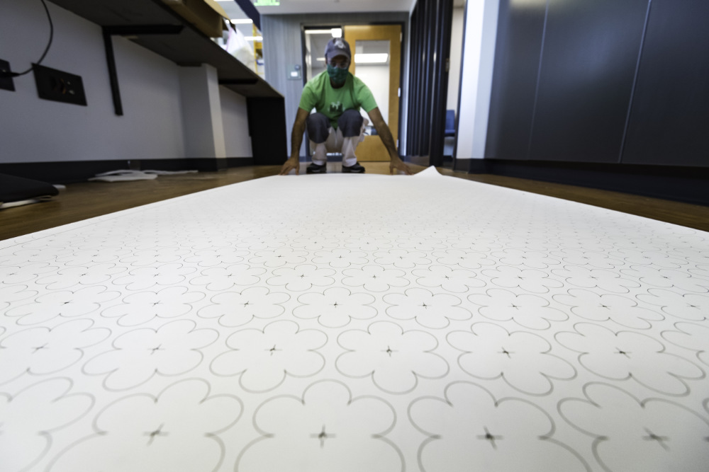 Man in green shirt rolls large sheet of paper with floral pattern out on floor.