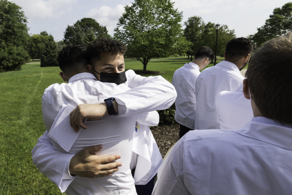 Two males in masks embrace outdoors surrounded by a few others.