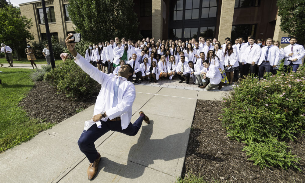Feature image Minhaj kneels and extends his arm to get a group photo of the class in their white coats.