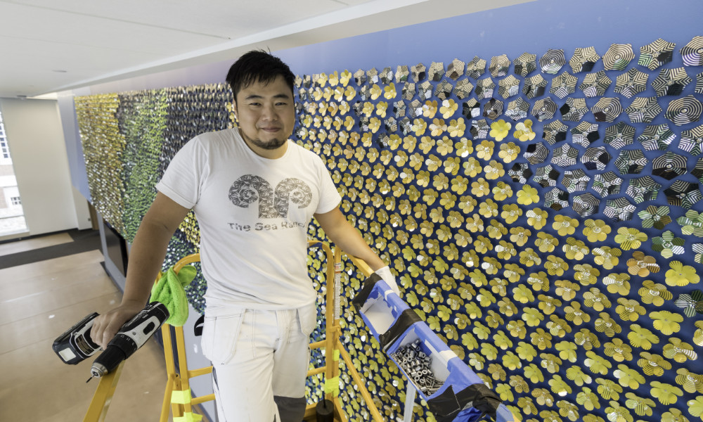 Jay Yan holds a drill atop a scaffold and smiles at the camera with his mosaic mural in the background.