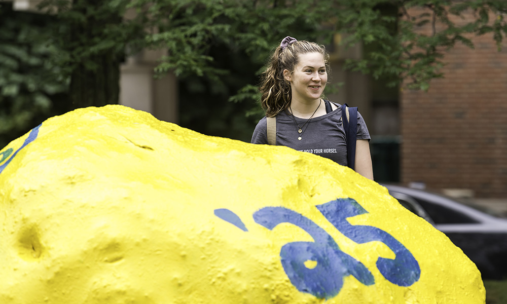 Feature image student standing beside a painted yellow rock with '25 painted on it