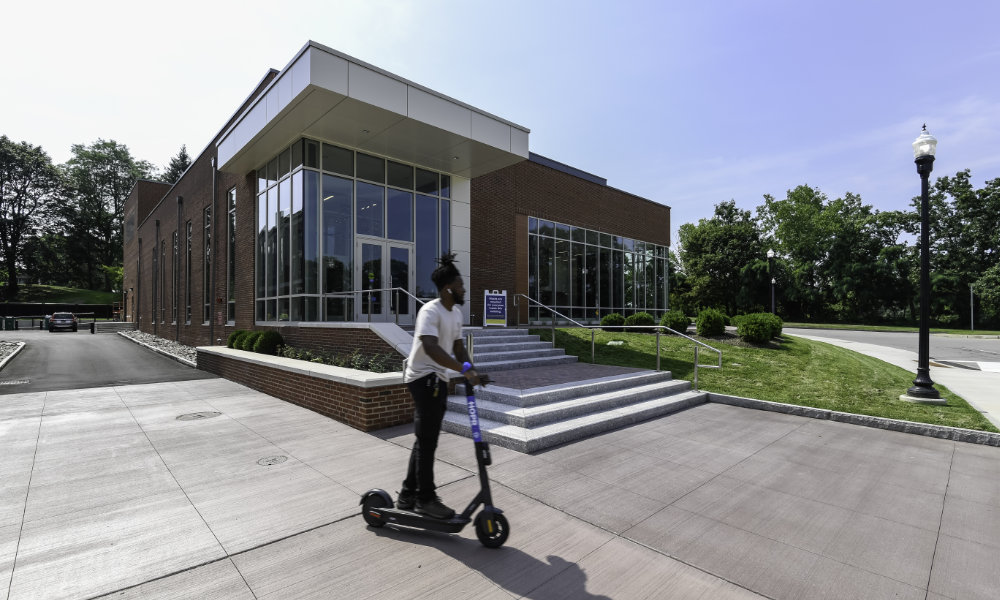 A man on a scooter rides past the exterior of the Sloan Performing Arts Center on a sunny day.