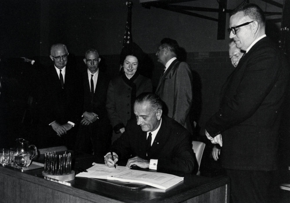 President Lyndon Johnson signs act with officials looking on.