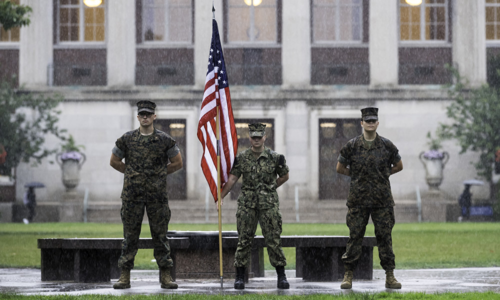 Feature image Three midshipmen in uniform stand on the Eastman Quad in the rain. The one in the center holds an American flag.