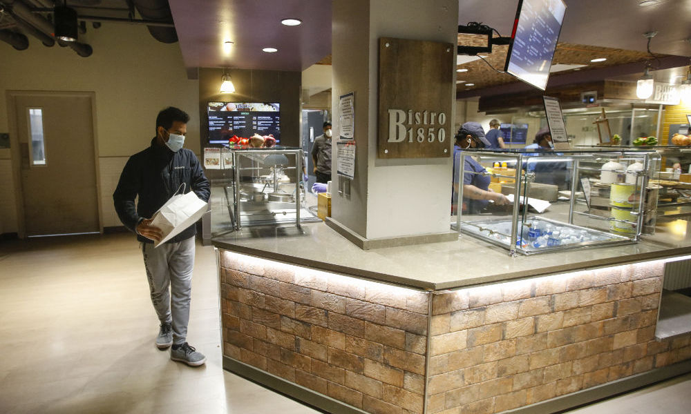 Feature image Student wears a mask and carries a to-go bag lunch at a food serving station.