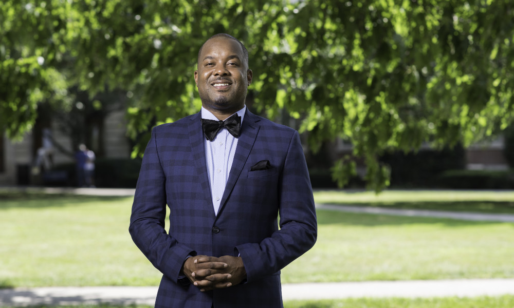 Jeffrey McCune in a suit and bowtie smiles at the camera outside on the Eastman Quad.