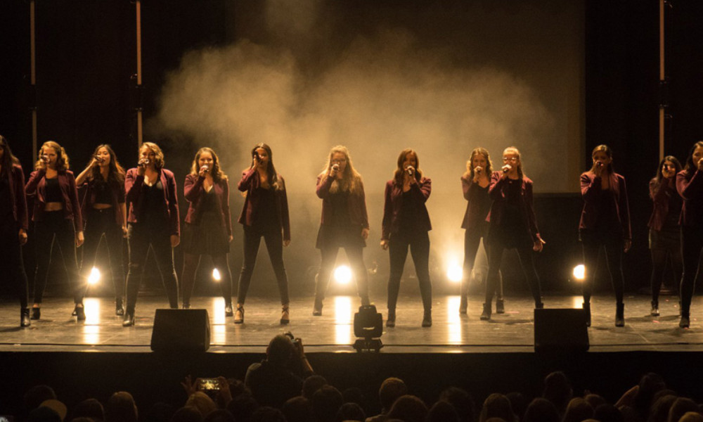 Feature image A dozen or so members of the all-female Vocal Point a cappella group lit up on a dark stage.