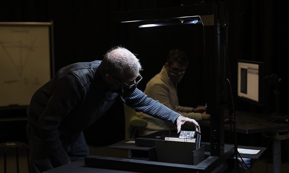 Man adjusts a book in the foreground with man seated at computer in the background