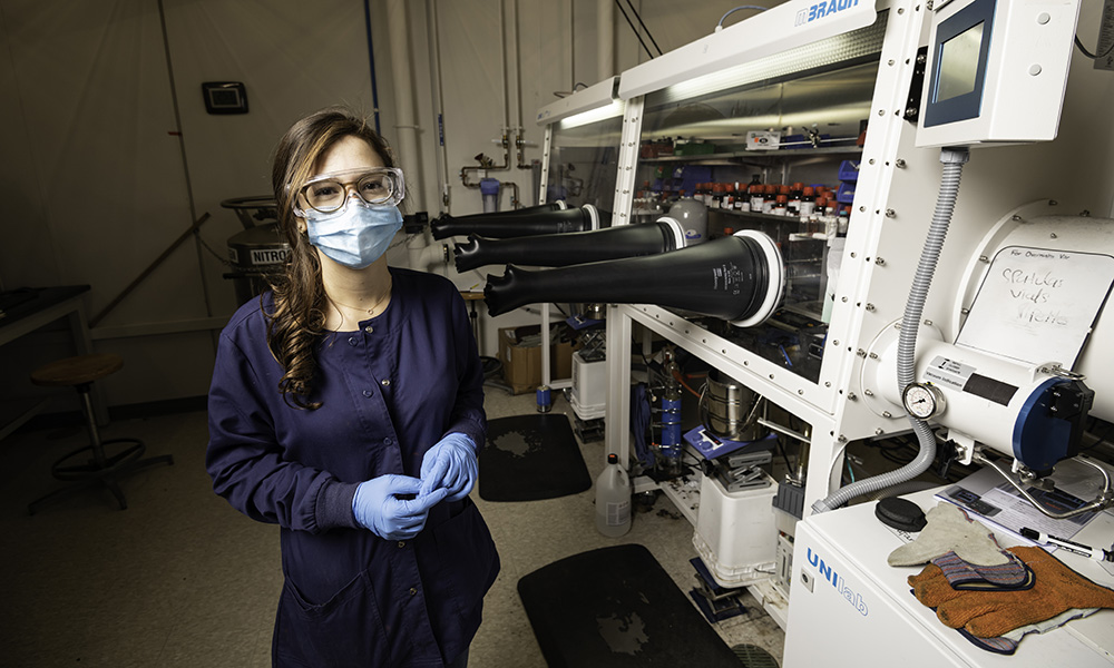 masked and gloved young woman stands in laboratory.