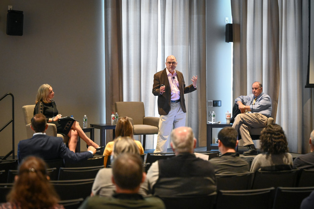 In front of an audience on stage is Sevin Yeltekin seated, Narayana Kocherlakota standing, and Clifford Smith seated.