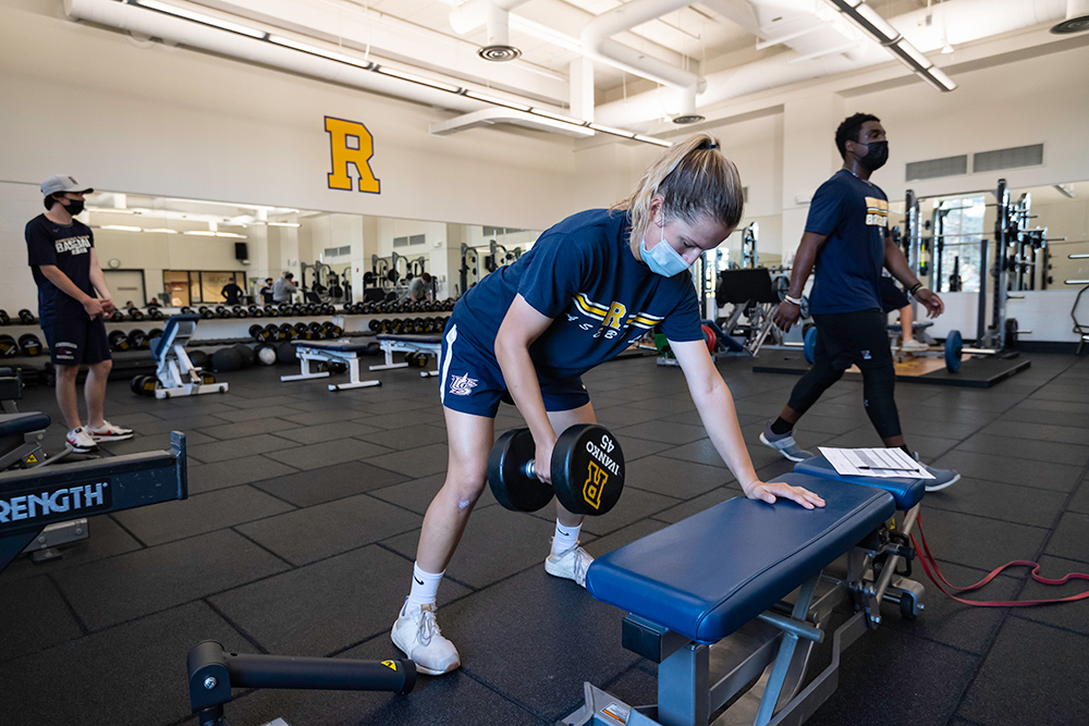 Beth Greenwood lifting weights on a weight bench with other baseball players working out around her.