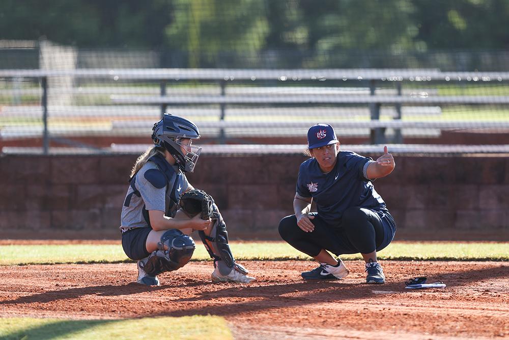 Beth Greenwood and Veronica Alvarez crouch at home plate as Alvarez points to the action on the field.