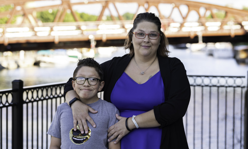 A mother and her son, who has FASD, pose for a photo near the Erie Canal with a bridge in the background.