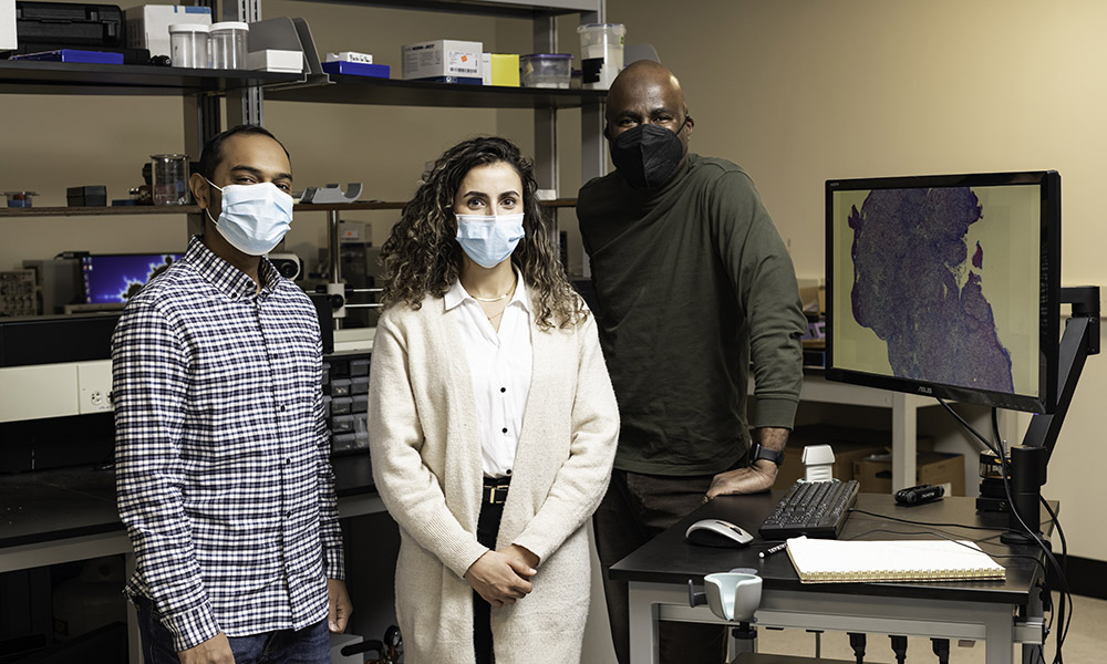 Three researchers stand in a lab beside an image of a tumor.