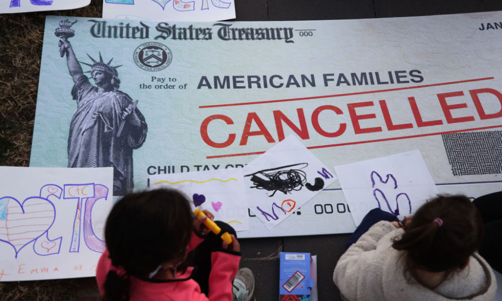 Feature image Two children seem from behind create drawings to accompany a giant stimulus check made out to American families with "cancelled" stamped across it.