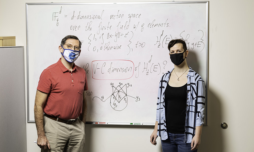 Professor and student standing in front of smartboard with equations.