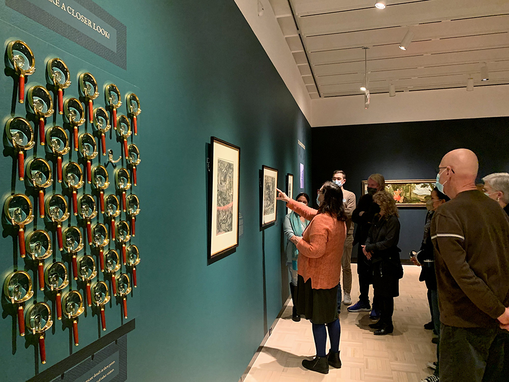 A museum wall features magnifying glasses hung on hooks, with a small group of people using them to look at the prints on the wall. 